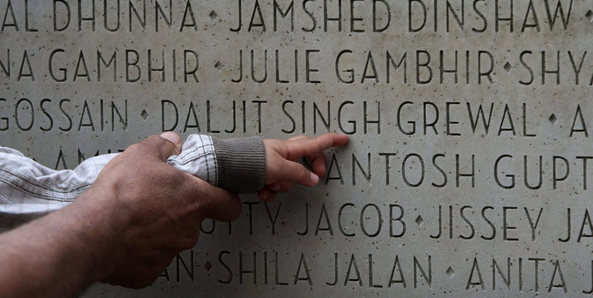 An adult’s hand holds a child’s wrist as the child points toward the name Daljit Singh Grewal on a stone memorial with hundreds of names.