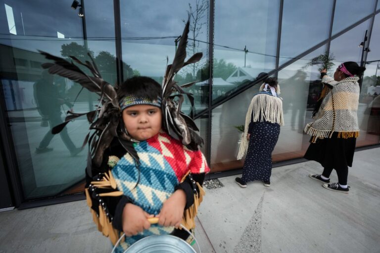 The boy is in regalia that includes a feather headdress. Behind him two people face the glass panels of the tower.