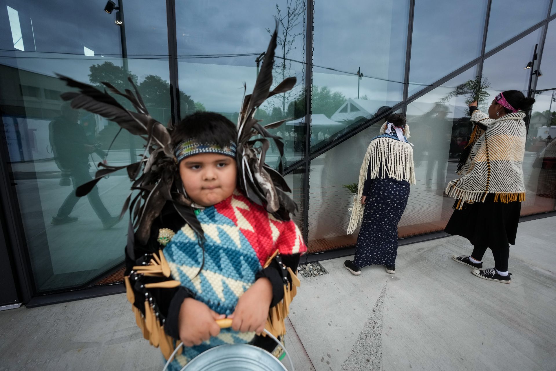 The boy is in regalia that includes a feather headdress. Behind him two people face the glass panels of the tower.