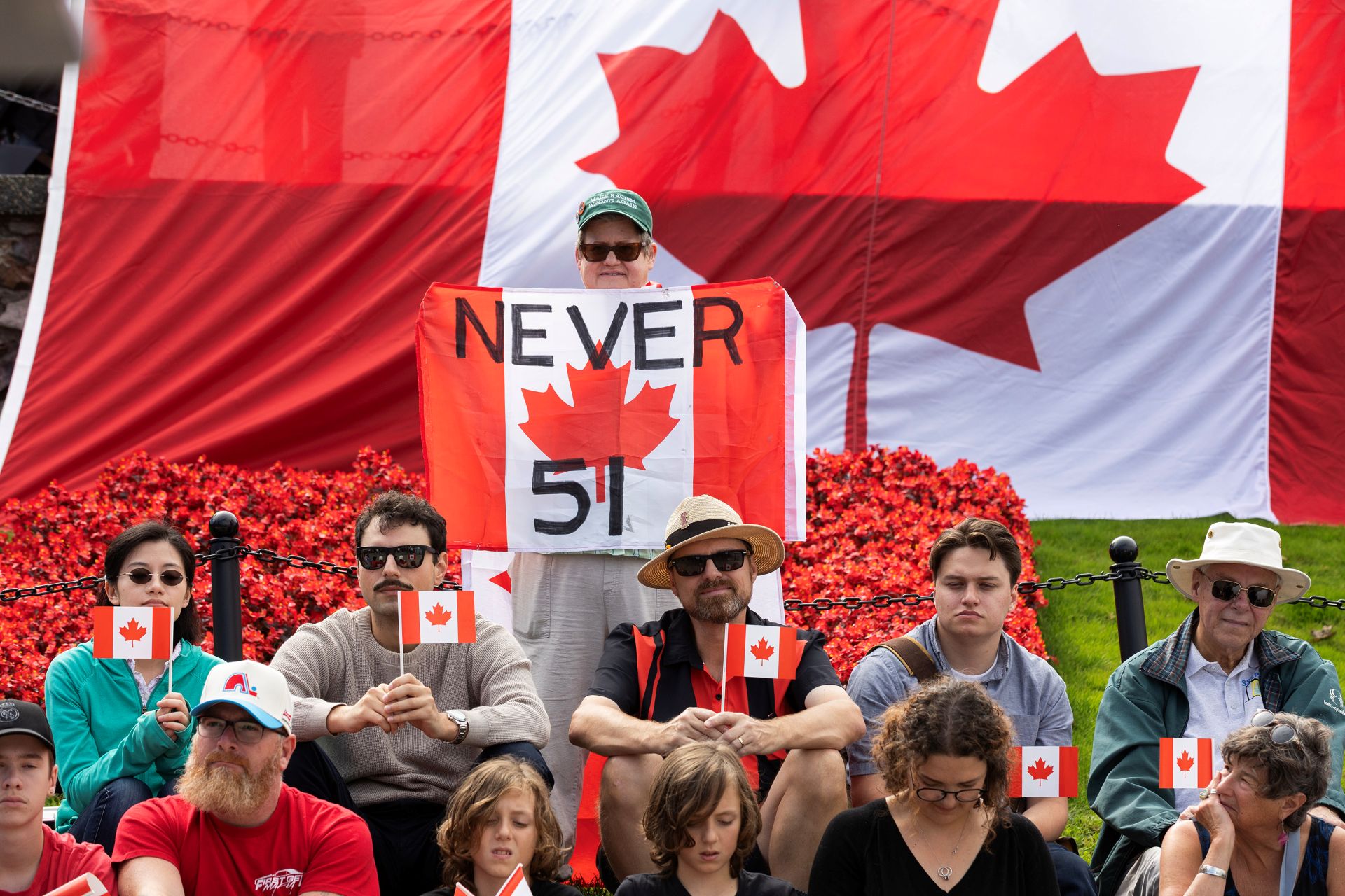 People holding small Canadian flags and a sign saying “never 51st” sit in front of a larger Canadian flag.