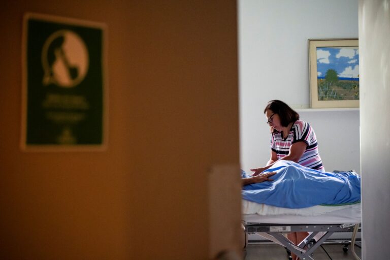Une porte entrouverte laisse voir une personne s’occupant d’un patient allongé dans un lit d’hôpital, dans une chambre où la lumière entre par une fenêtre.