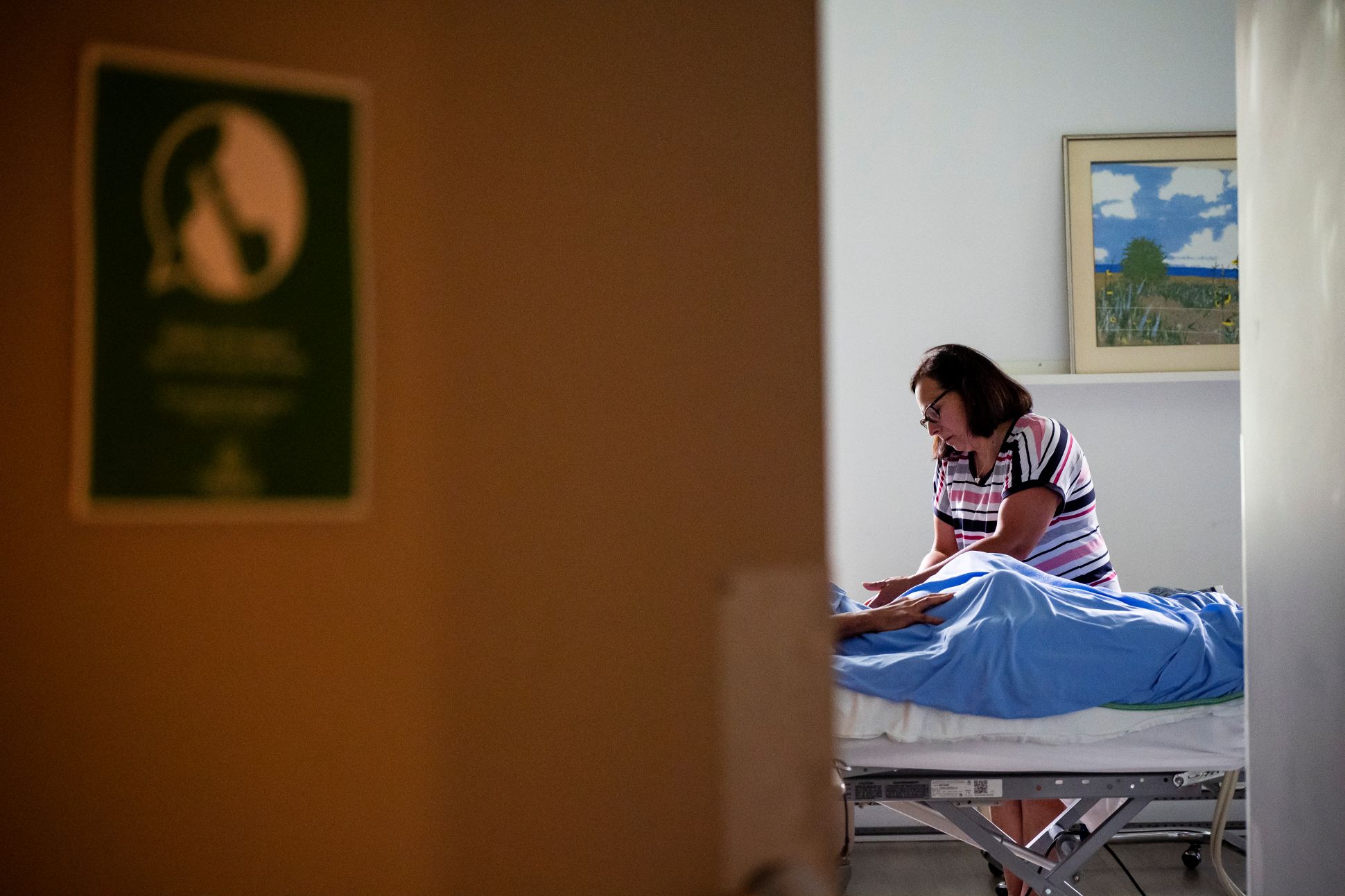 Une porte entrouverte laisse voir une personne s’occupant d’un patient allongé dans un lit d’hôpital, dans une chambre où la lumière entre par une fenêtre.