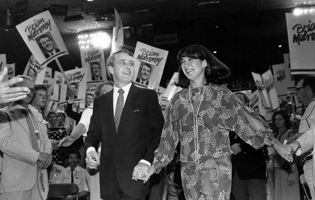 A black and white photo of Mila and Brian Mulroney at a political convention surrounded by supporters with signs. 