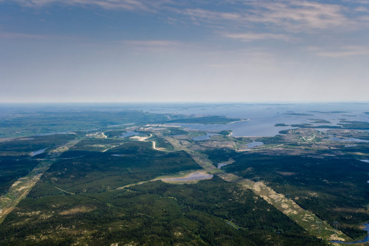 Aerial view of a wide body of water in the distance with dense forests and strips of land cleared for hydro poles and wires cutting across the landscape.