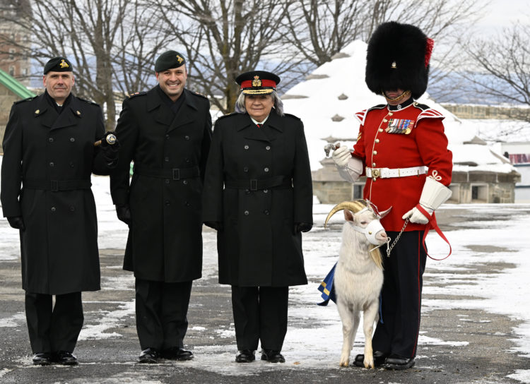 A smiling Simon is flanked by three men and a goat. On one side are two soldiers in dark berets, black coats, black pants and black shoes. One of the men is smiling, one is not. On Simon’s other side is a man in regalia: tall, fluffy black hat, red jacket with ornate shoulder pads, a row of medals pinned to the breast, a white parade belt, long-armed white gloves, black pants with a red side stripe, and black boots. The man is holding an ornamental cane in one hand; in the other he holds a short chain connected to Batisse, the long-haired white Persian goat, who is sporting a white muzzle, golden covers for his horns and a blue and gold fabric hung over his back. The ground is covered in patches of snow.