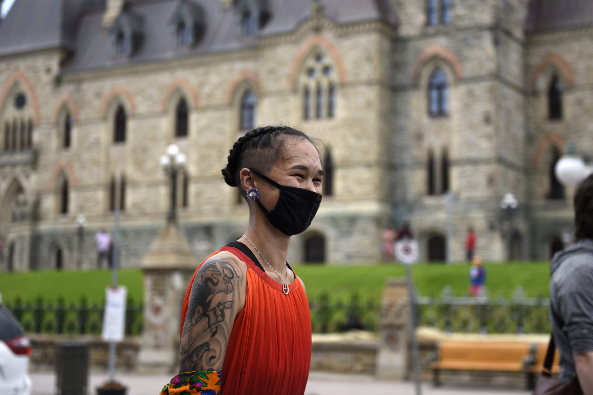Qaqqaq is wearing a black mask. Her hear is shaved on the side and braided tightly on the rest of her head. She is wearing an orange sleeveless top showing tattoos covering all of her right arm. It looks like she is smiling under the mask.