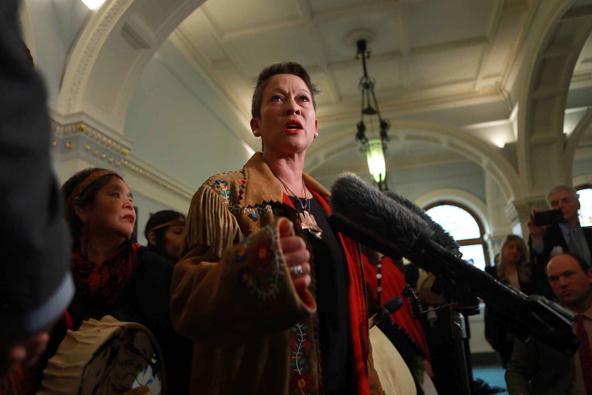 Mark speaks into a cluster of microphones in an elegant room with coffered ceilings and archways. She is surrounded by Indigenous supporters. She holds a flat, wide drum at her hip and is dressed in a tan rawhide jacket with tassels and green, pink and red flowers painted on the cuff paired with a black shirt. She’s got a bronze ornament on a beaded chain around her neck.