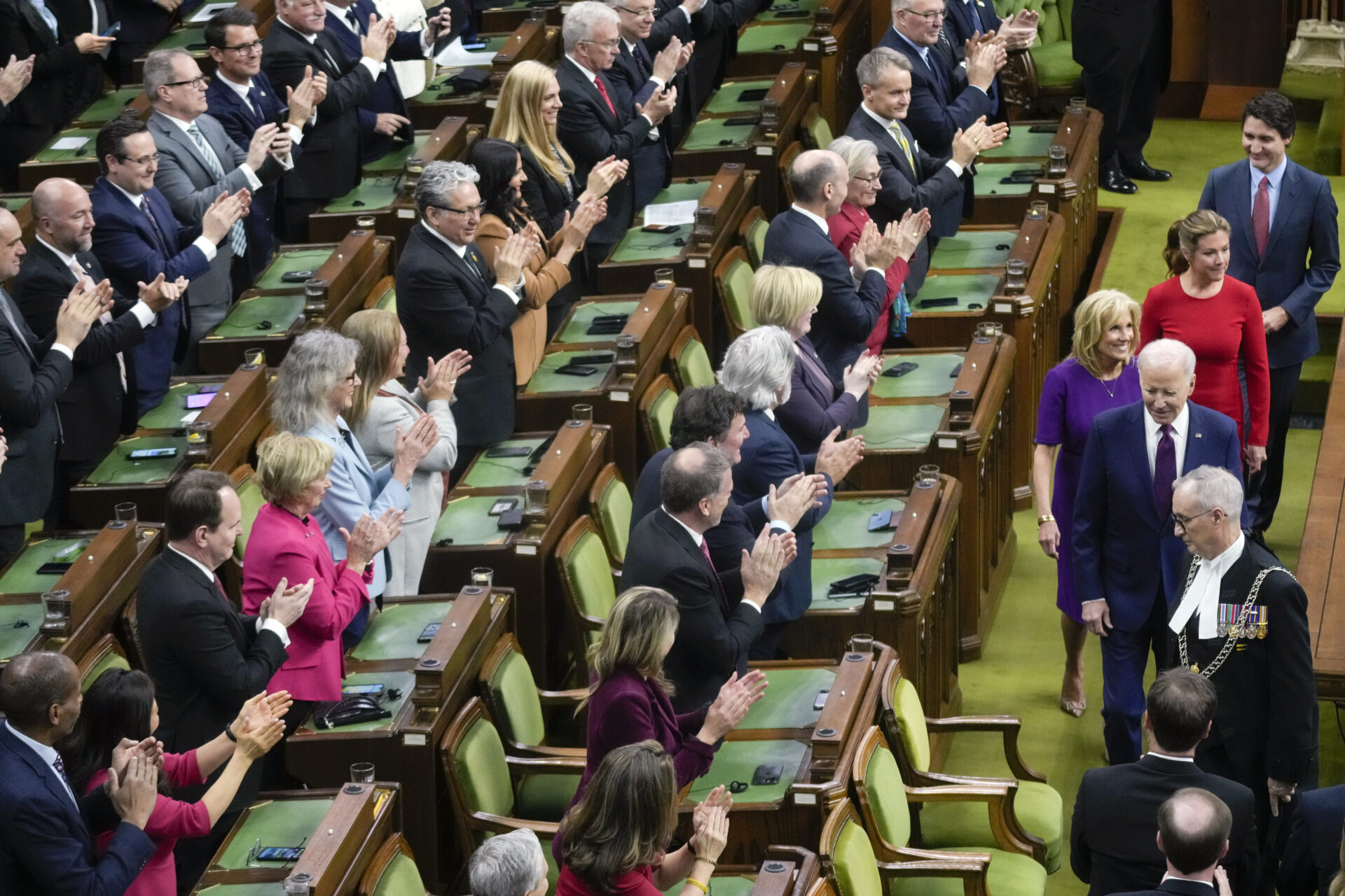 Four rows of MPs stand in their seats and applaud as Biden, Trudeau and their wives pass in front of them. The MPs are a mix of women and men, with the men in dark suits and the women wearing jackets in various colours, some pink, burgundy, powder blue. The floor is covered in an olive green carpet. The desks in front of each MP are wood with square green blotters. Each MP’s chair is wood with olive-green velvet backing and cushions. Most everyone is smiling. 