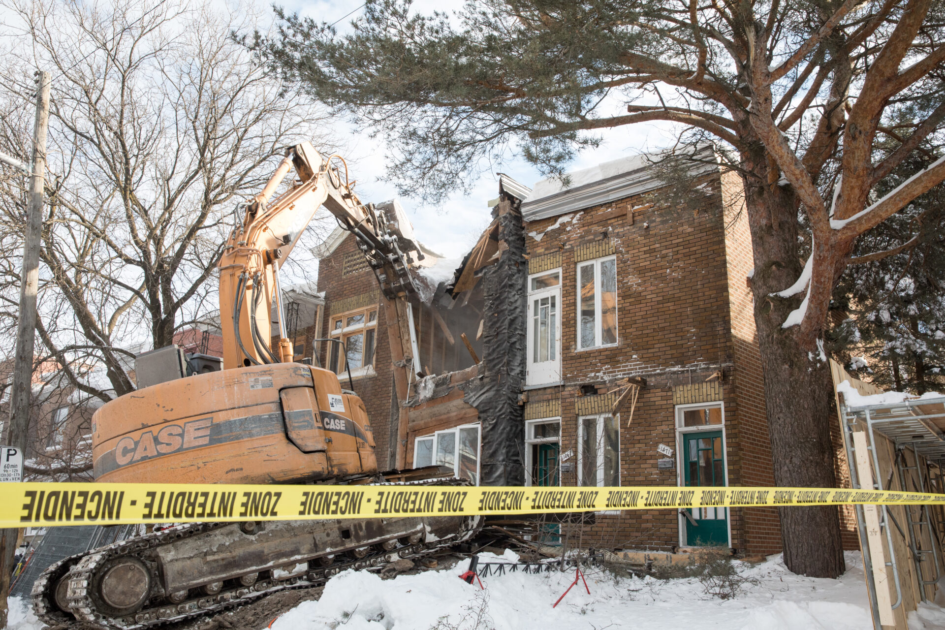 The claw of an excavator slashes through the roof of a two-storey brick fourplex on a tree-lined street. The front lawn is covered in snow.