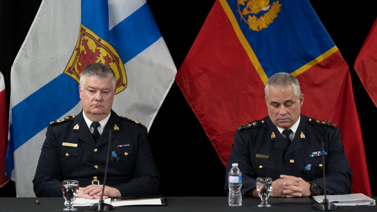 Both men sit at a long table covered in a black tablecloth. They are both wearing black uniforms with pins signifying various honours and achievements. Behind them are two large flags, one the Nova Scotia flag, the other the flag of the RCMP. 