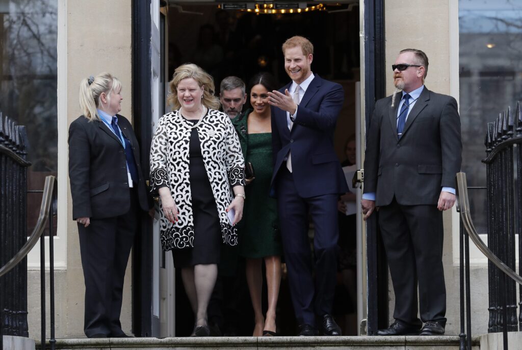 Charette and the royals are part of a group of four people coming out of an entrance flanked by a man and woman holding the doors open. She is smiling, and her hair moves freely with the wind. She’s in a black dress and a long white cocktail jacket that hangs open. It has a busy pattern in black and white. The duchess is in an emerald green dress and short jacket. The prince is in a black suit. Both royals are smiling.