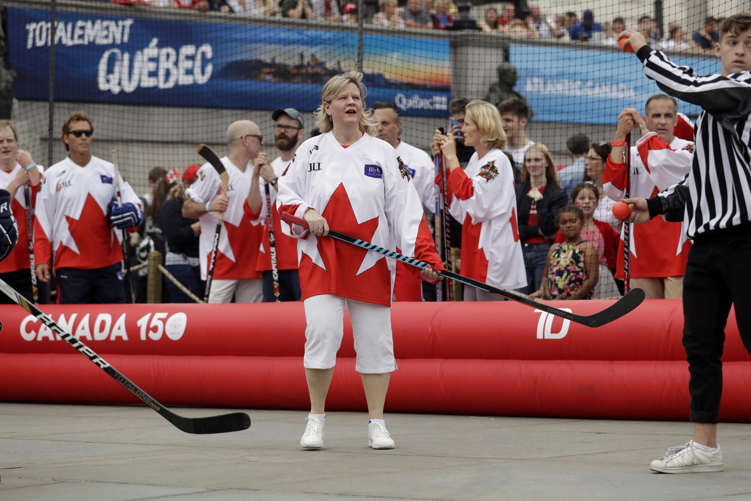 Charette is standing in an inflatable “rink” whose border is red and says “Canada 150” on it. She and her teammates are dressed in red and white jerseys with a large maple leaf motif. Her calf-length pants are white, and her sneakers are white. She’s holding a hockey stick. Many spectators sit in bleachers in the background.