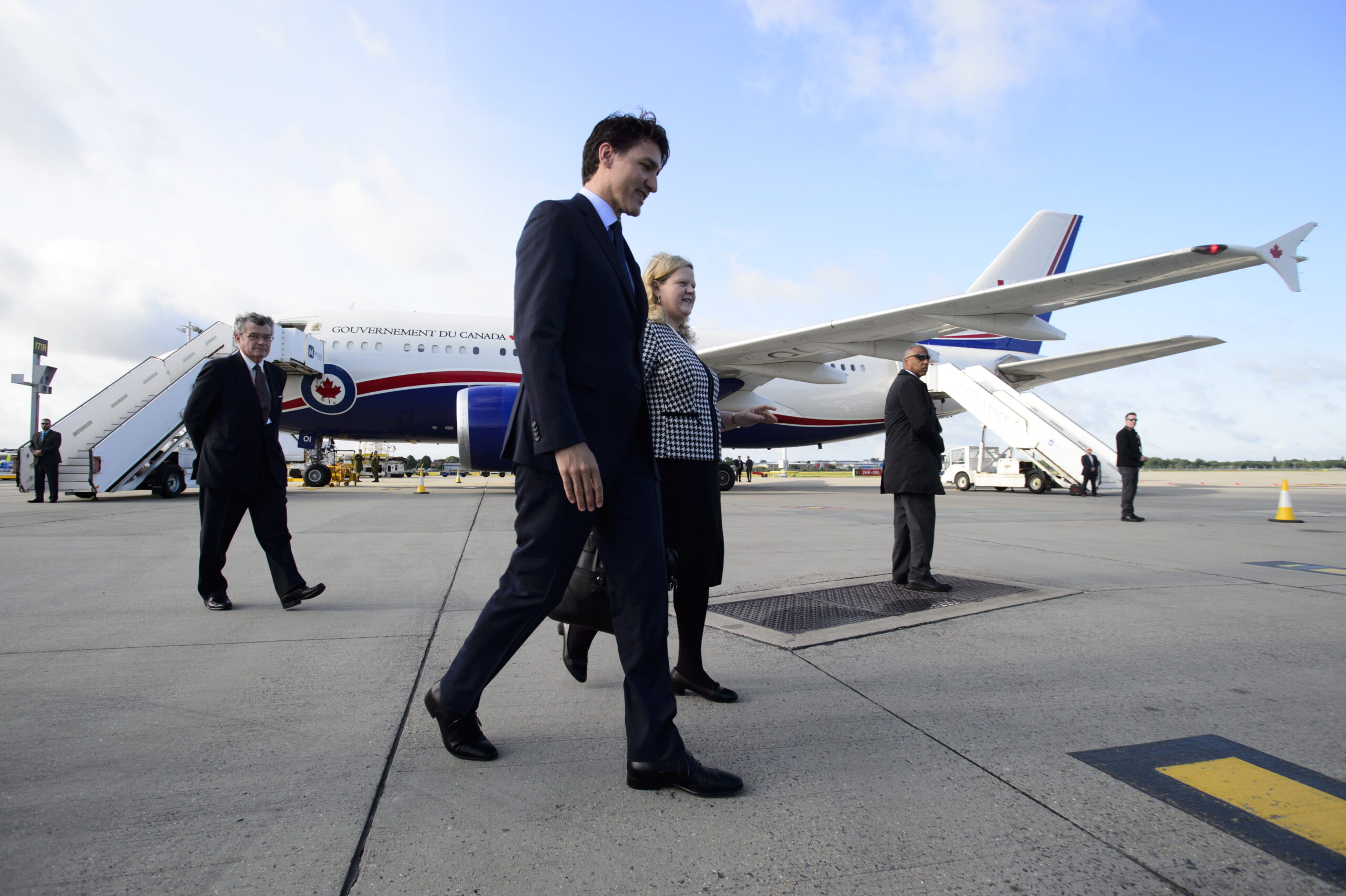 Charette and Trudeau walk side by side, with four men positioned variously around the tarmac with the government jet in the distance, its two stairway ramps in use at the front and back of the plane.