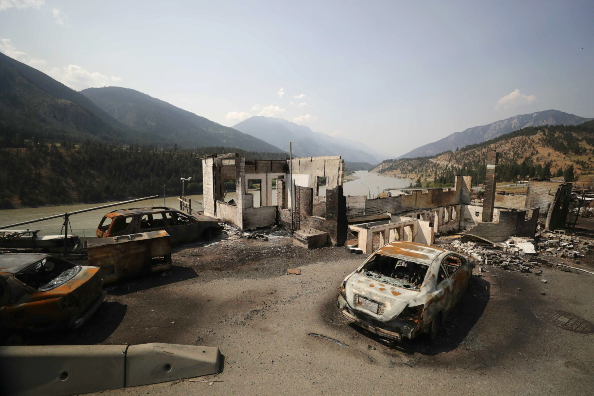 Three burned-out cars sit among the remains of homes in a community along the shores of a river, with mountains on the other side and in the distance. Only the foundations and concrete walls of the homes remain.