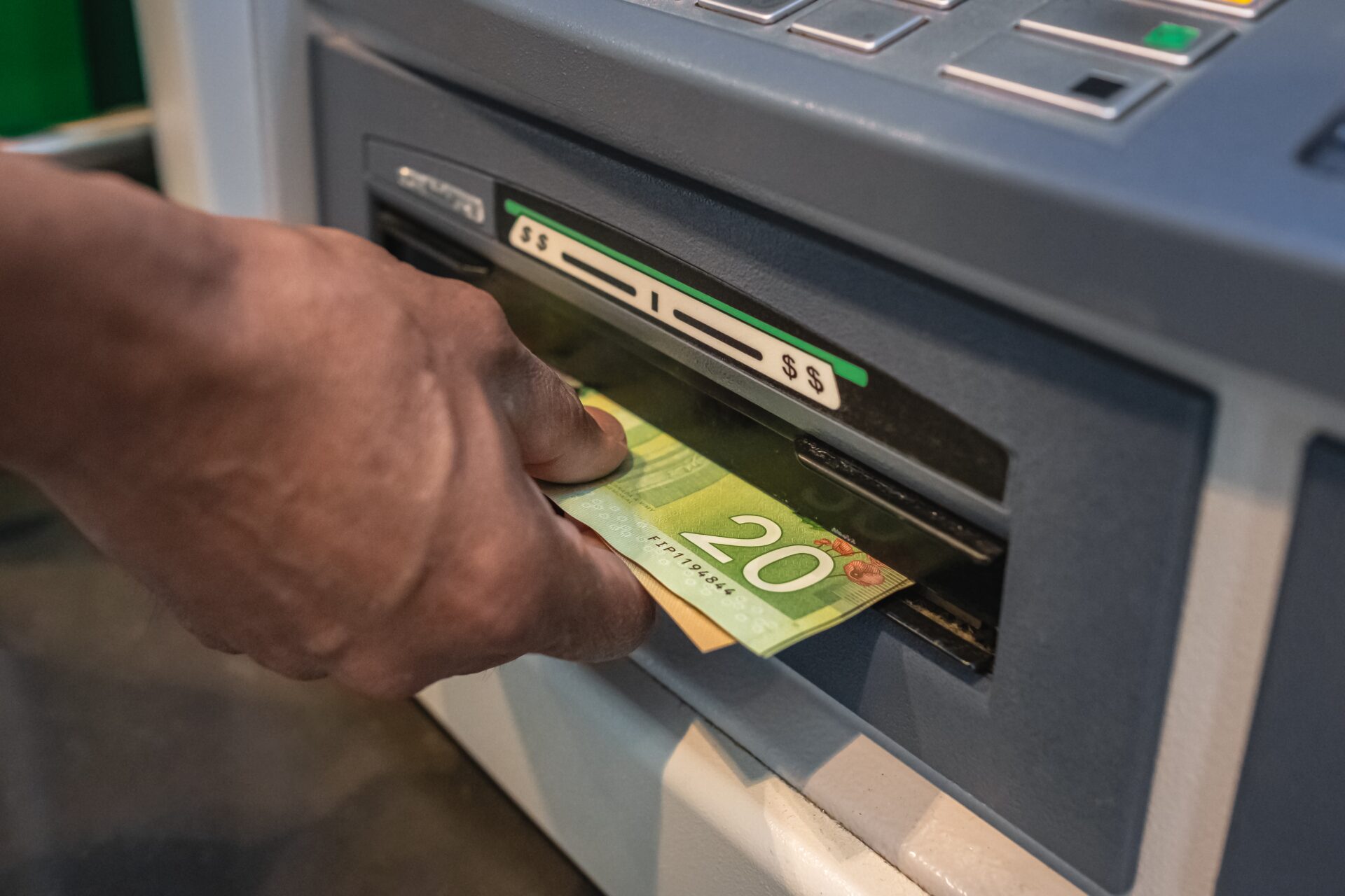 Close-up of a money slot at a cash-dispensing machine, with a man’s hand taking a slim stack of $20 bills as it comes out.