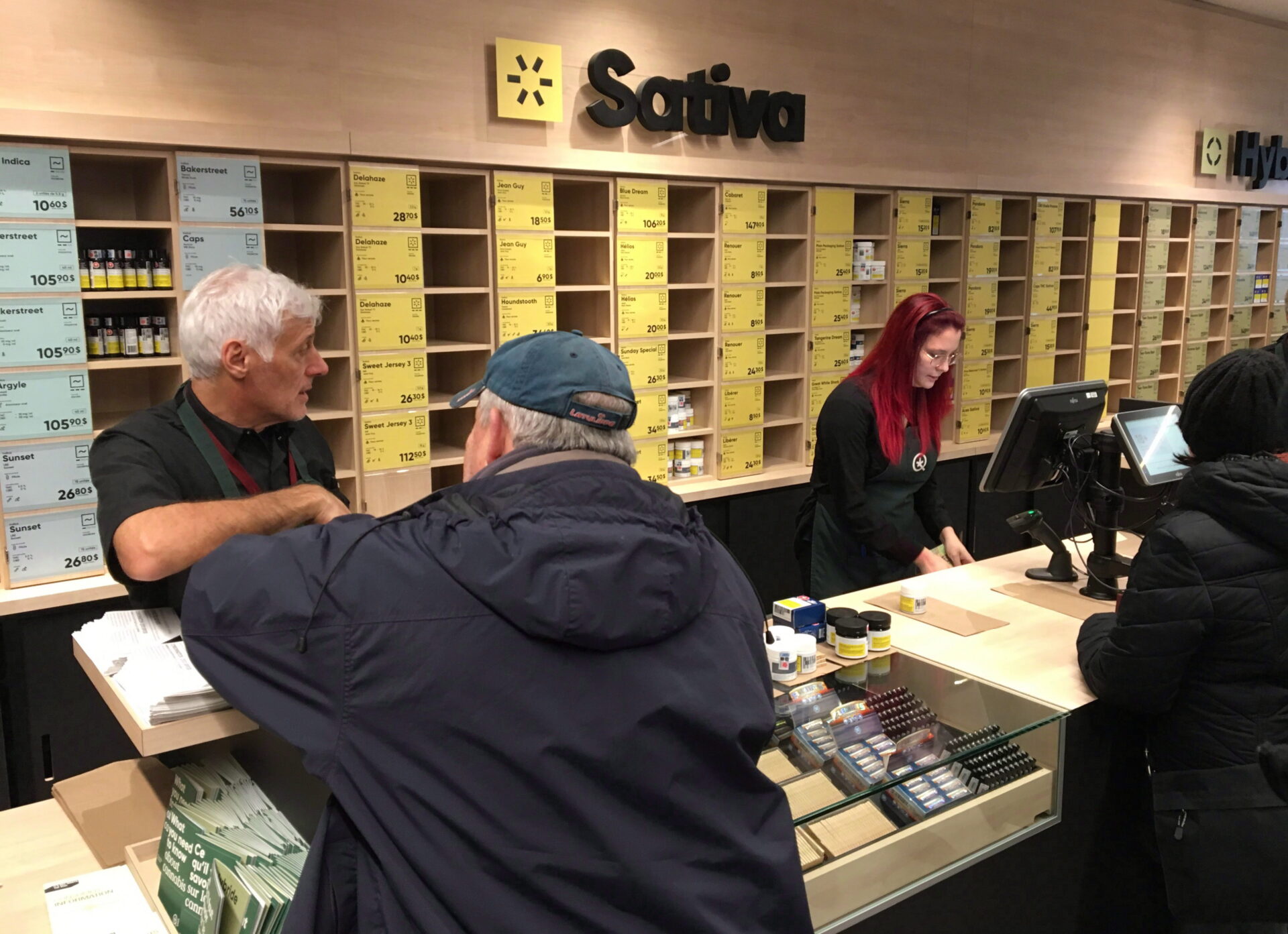 Three customers stand at a long counter where two employees serve them behind computers. A small selection of products is stocked on the counter. The back wall is filled with cubby holes that hold small products. Many of the holes are empty. Above the cubby holes is the word “sativa” in large black letters.