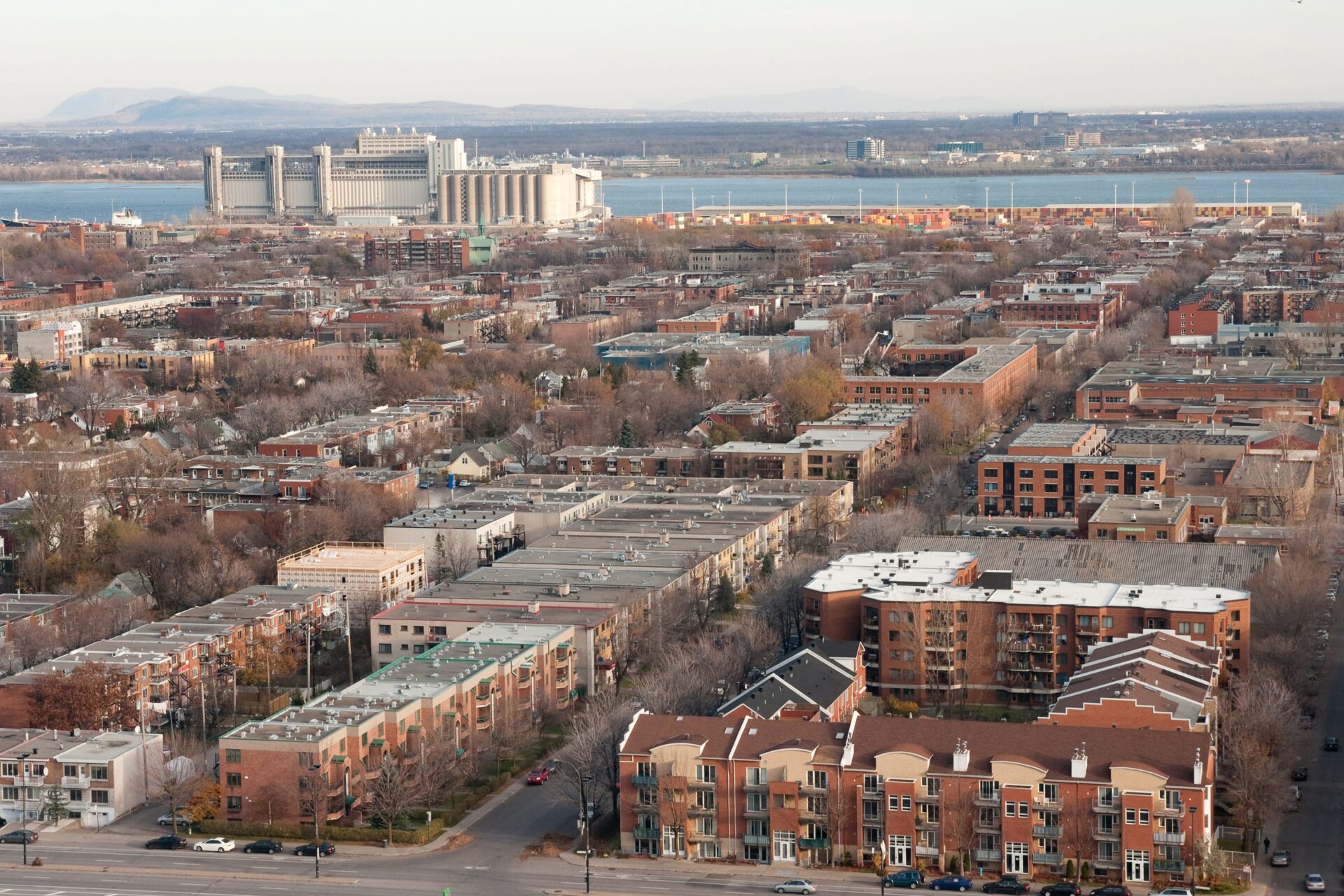 An aerial view of the streets of the neighbourhood with the St. Lawrence River in the distance. The area is characterized by brick rowhouses and plexes three and four storeys high, with mature trees lining most streets.