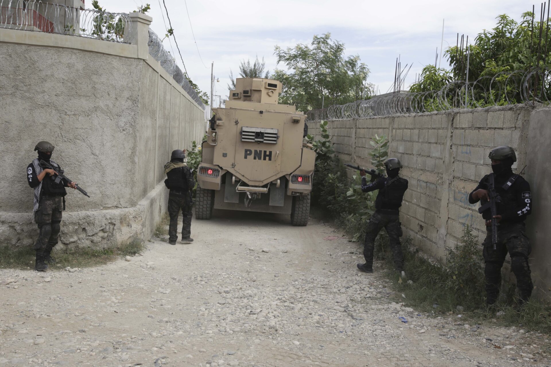 Four officers dressed in black jackets, black helmets and dark fatigue-patterned pants are positioned along a narrow dirt road that has stone walls on both sides. They carry rifles. One of them is aiming it toward a tan-coloured large, boxy police vehicle that is a few feet down the road. Barbed wire sits on top of the walls.