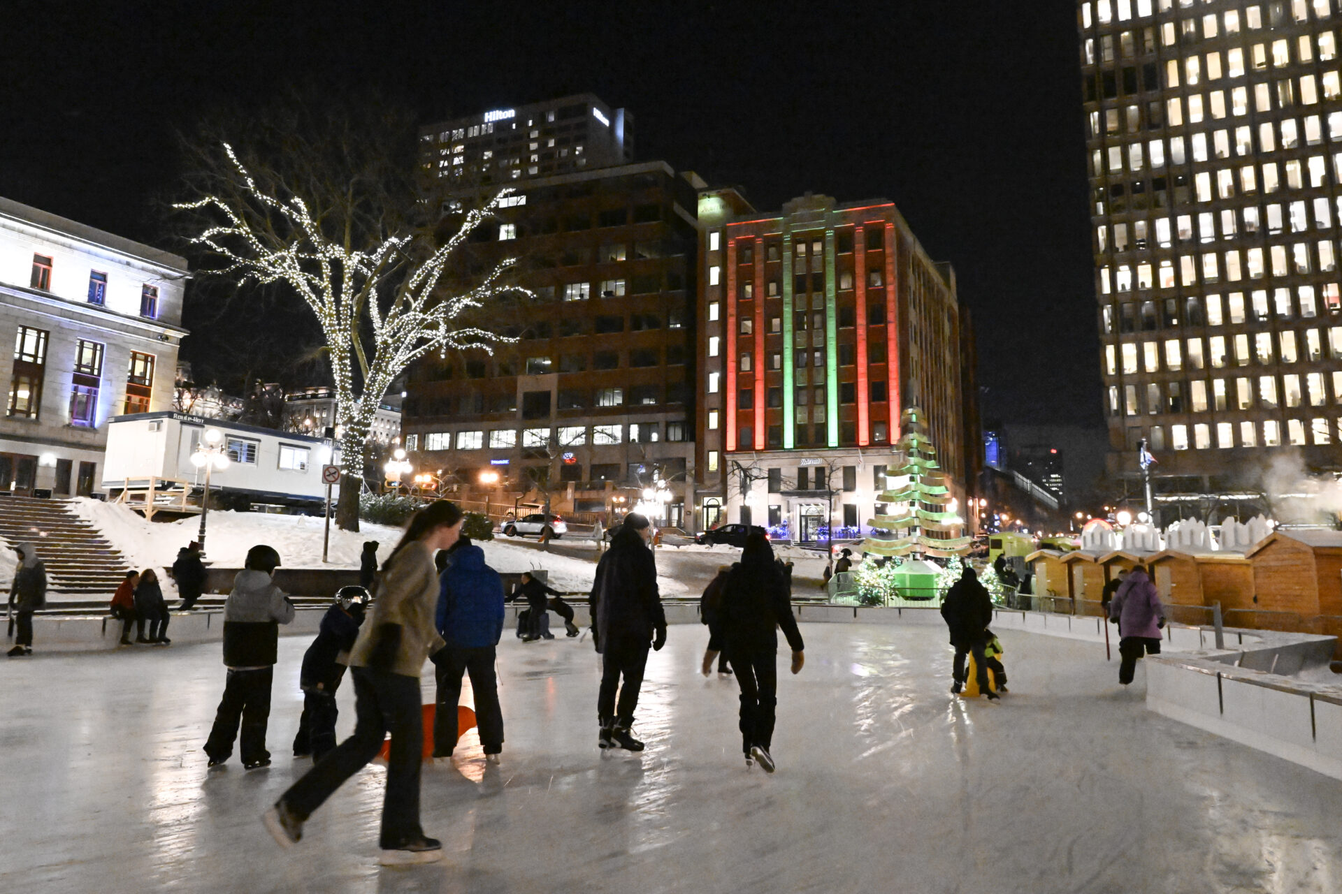 Ten or 12 people skate on ice surrounded by three buildings of varying size. One is a tower, another is several storeys high. One is decorated in red and green lights, and a tree is lit with white lights.