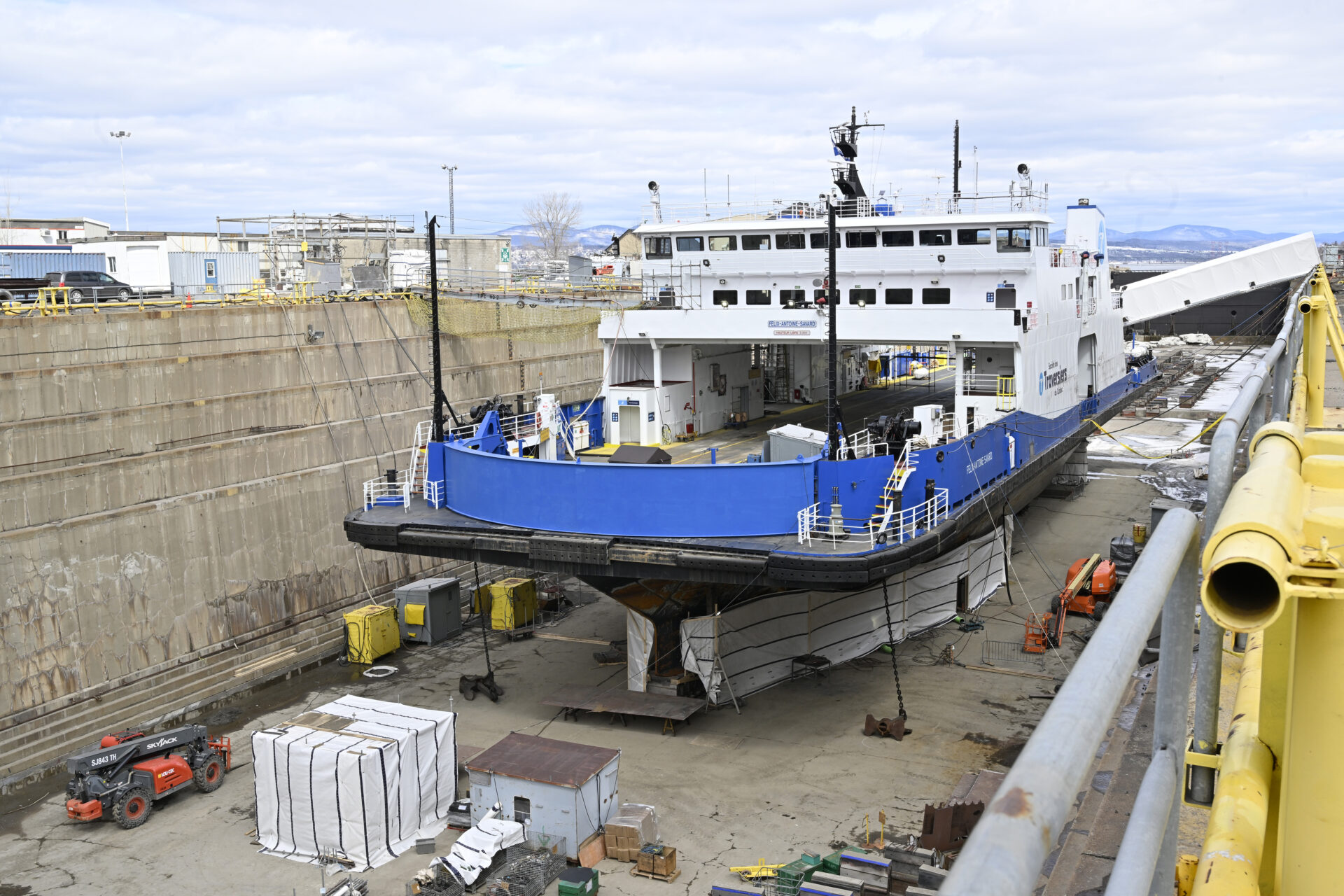 The ferry is long, wide and shallow, with a blue metal hull, a black bumper that runs its length at its waterline. It is sitting in a concrete enclosure that has been emptied of water It is surrounded by small machinery and supplies.
