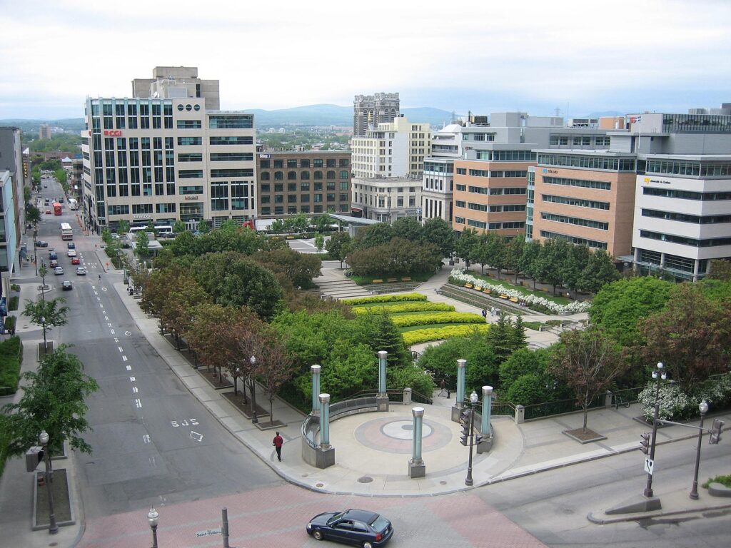 A full city block is taken up by the park, which is bordered by a thick cluster of trees of various varieties, some evergreen shrubs, some with reddish brown leaves. In the centre of the park are flower beds and sitting areas. It’s a tidy green space surrounded by mid-rise buildings. In the distance is a treed sector of the city and hills beyond them. 