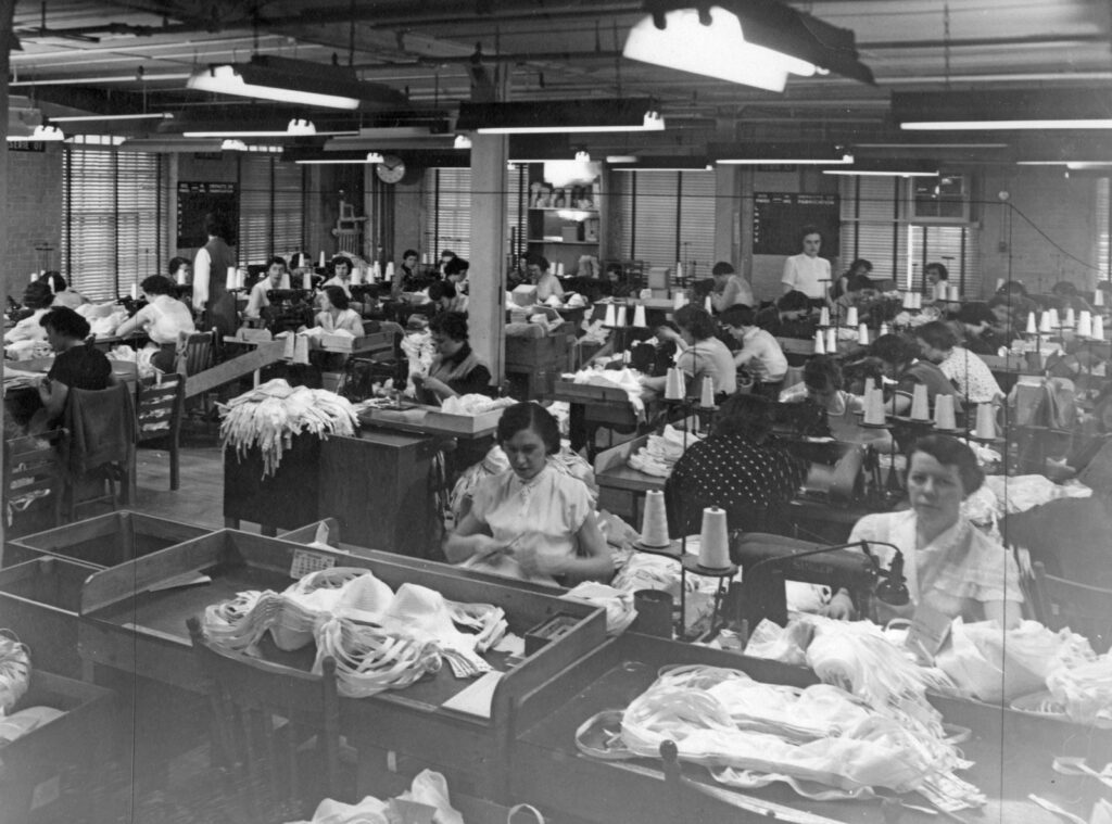 Rows of desks fill a room. At each desk is a woman working at a sewing machine. In front of them are small piles of newly created bras on desks. Blinds cover all the windows. It’s a black and white photo. 