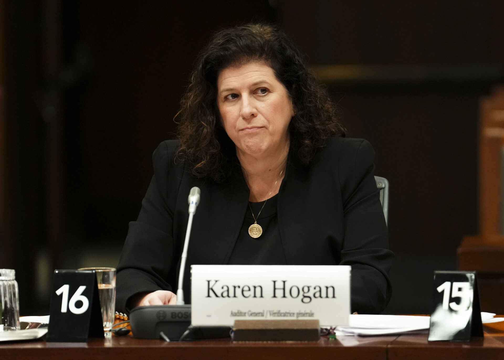 Mme Hogan est assise à une longue table en bois, avec un microphone et un badge devant elle. Elle porte un blazer et une chemise noirs et écoute d’un air sérieux. 