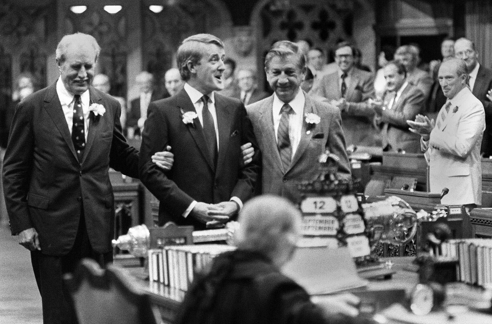 Rows of men in suits stand smiling and clapping as they watch Mulroney, who is flanked by two smiling men in suits. Each has an arm through Mulroney’s.