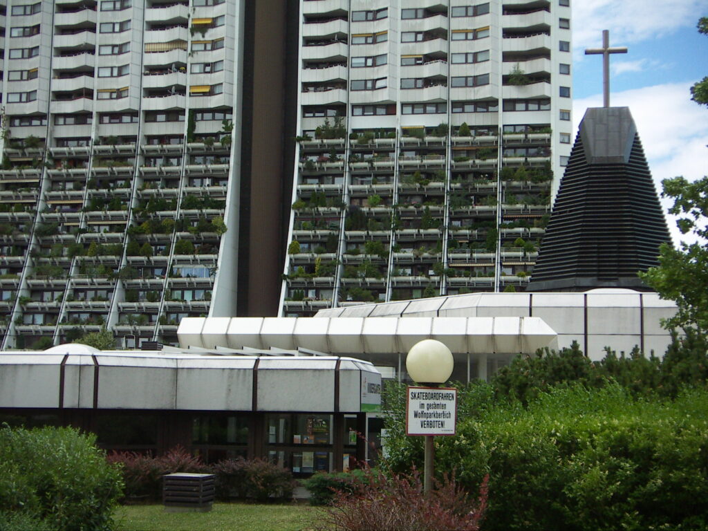 About 17 storeys of this high-rise building are shown. There is a ground-floor concourse entry surrounded by tall and small shrubbery. The shape of the building is wide on the lower floors, tapering to a thinner structure for the higher floors. The first 10 or so floors have balconies that are stuffed with greenery. The upper balconies are more angular, jutting out on one side. 