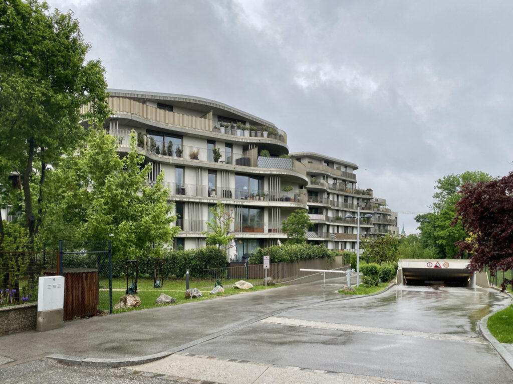 Two of the buildings are shown. They are five storeys high, and each floor has a balcony/terrasse that is curvy. Each unit has floor-to-ceiling windows. Trees and shrubs surround the property and potted greenery is a fixture on most of the terrasses.