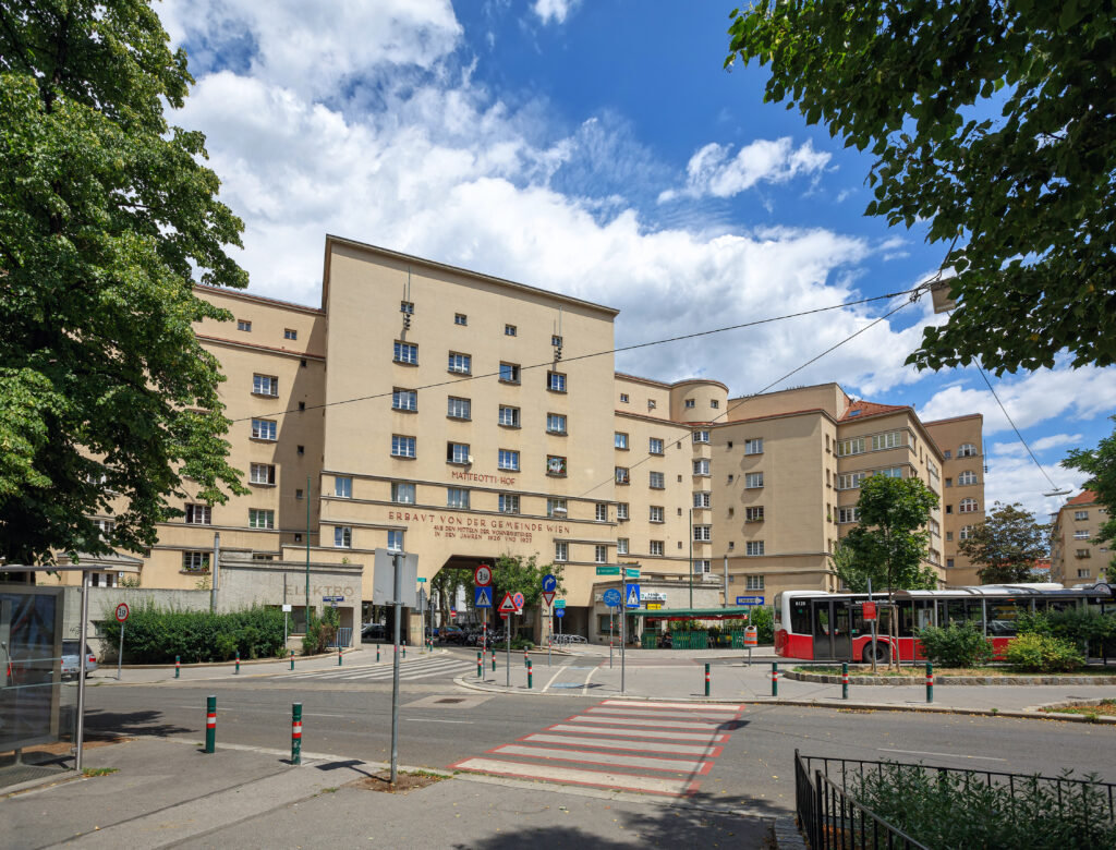 The building is about eight storeys high and has a handful of different parts to it. It is L-shaped with some wings extending out in a diagonal direction. Shrubs and trees are planted nearby. A cred and white city bus is stopped in front. A street runs through an archway in the building.