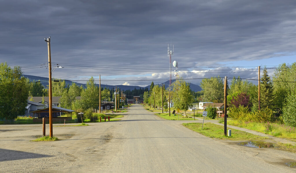 Bungalows and trees of various sizes line a road that makes up most of the photo. In the distance is a forest of evergreens, with mountains beyond.  