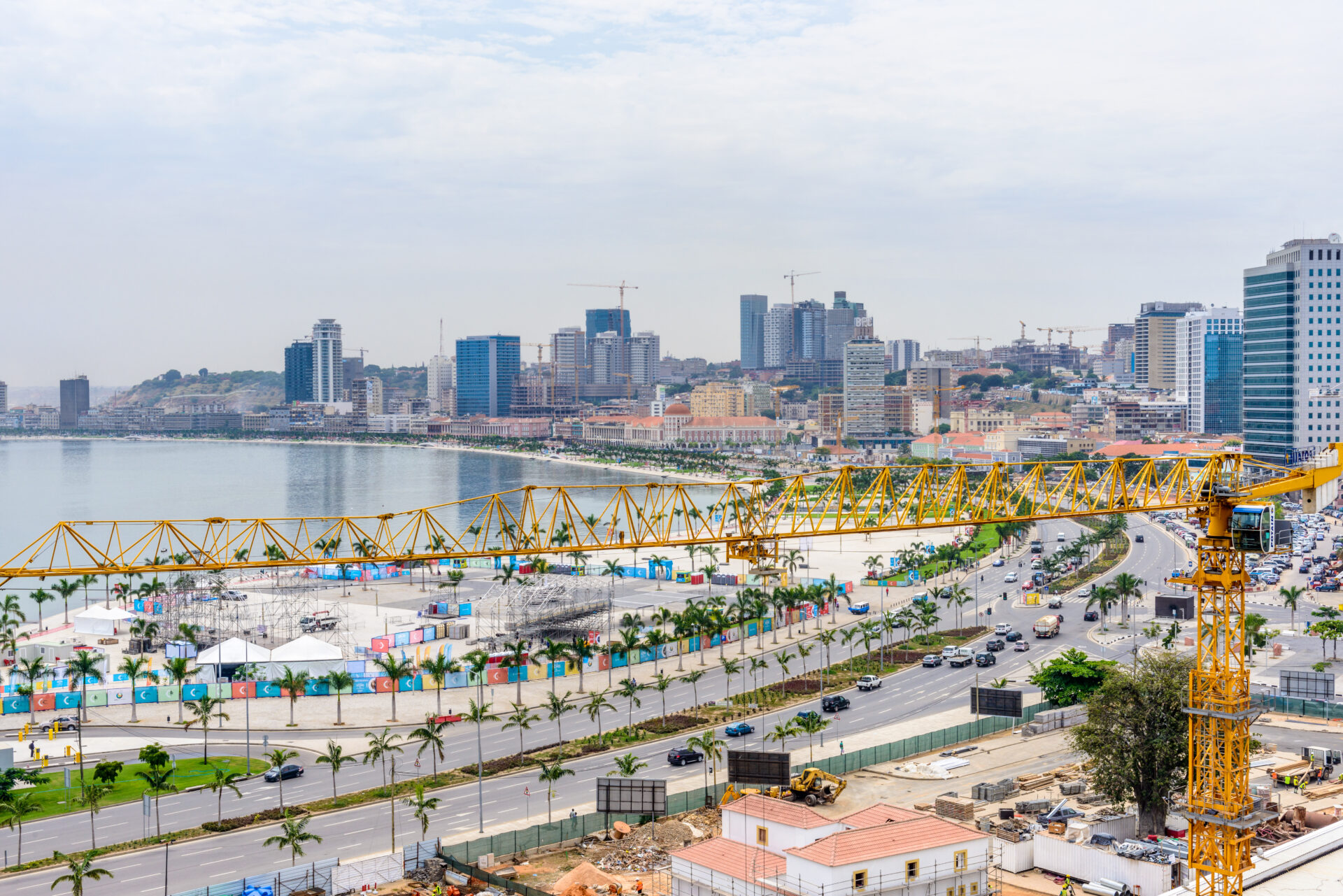 Construction cranes dot the skyline and foreground of a bustling city on the water, with a major highway lined with palm trees running through the downtown close to the shore. Lots of commercial towers. 