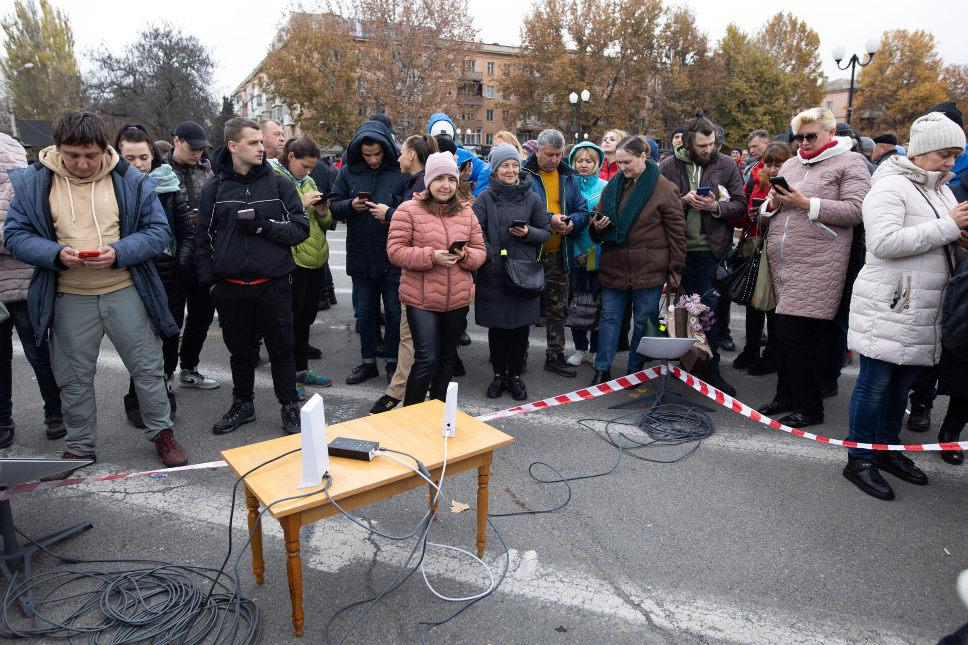 About two dozen people in winter coats stand around a small black metal box and two small white metal pieces of equipment sitting on a small light brown wooden table. The equipment is connected by grey cords to other equipment, all of it set on the pavement of an open area bounded by trees and a four storey brick apartment building. Everyone has a cell phone in hand.