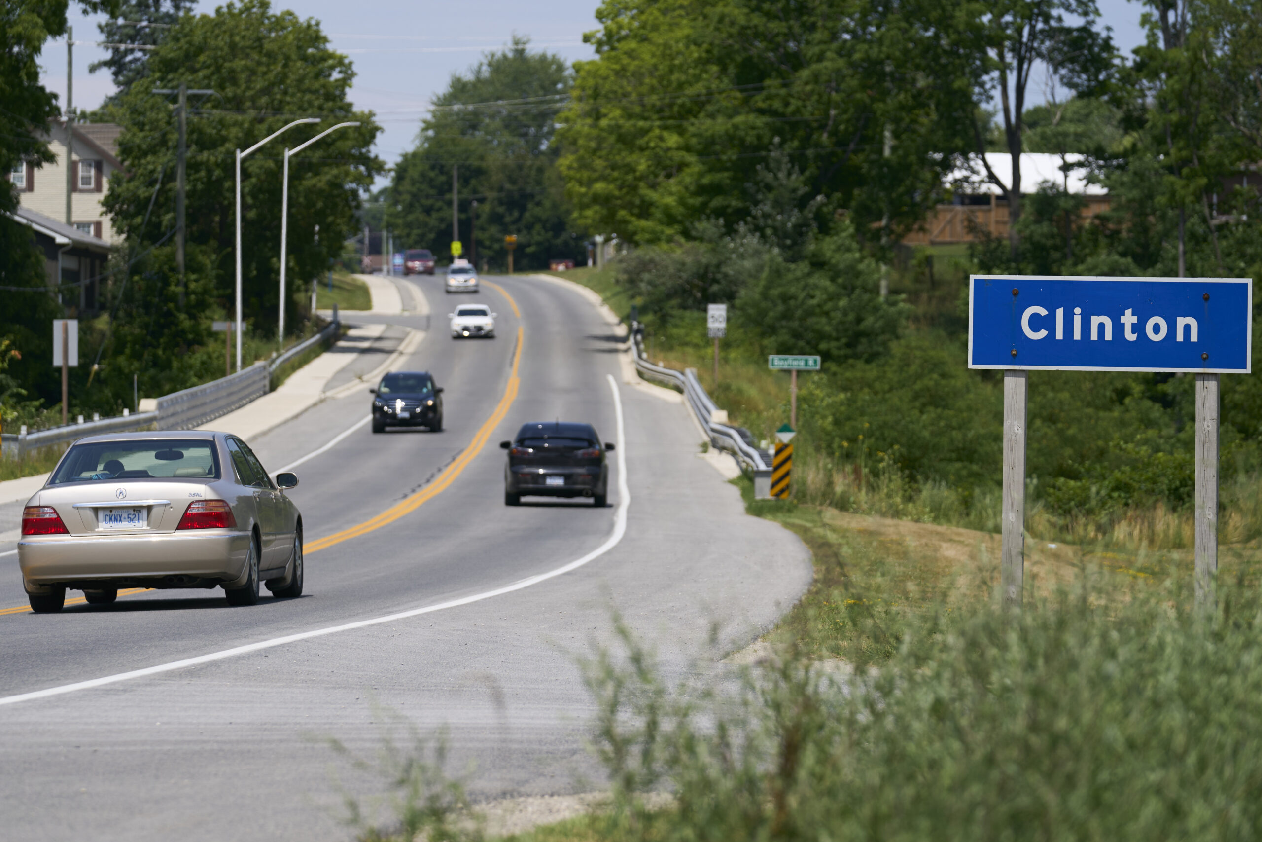 A single-lane highway with a handful of cars is lined with mature trees and homes.