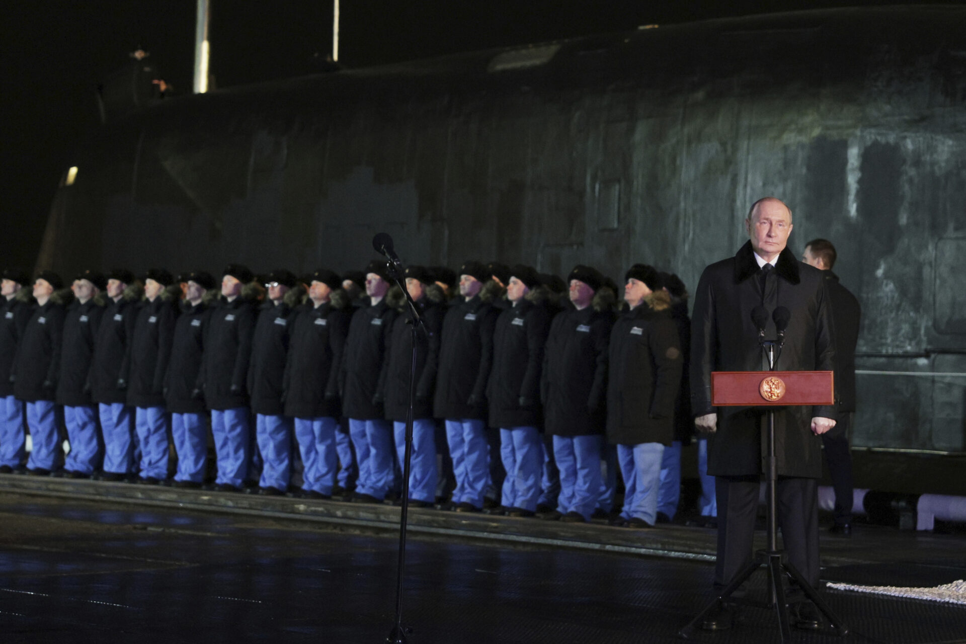 A dozen young men and women stand side by side at the edge of a dock. Behind them the submarine towers over them, its dark metal with a green hue in the night lights used for the ceremony. Putin stands off to the side at a podium.
