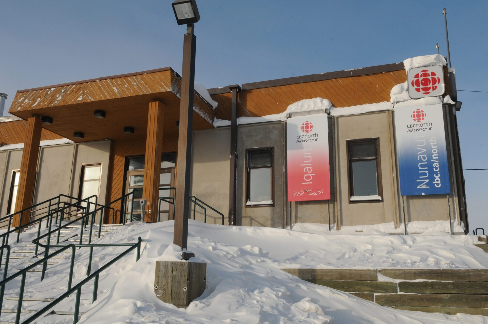 The one-storey building has front steps with a thin black metal railing. Snow drifts surround the building.