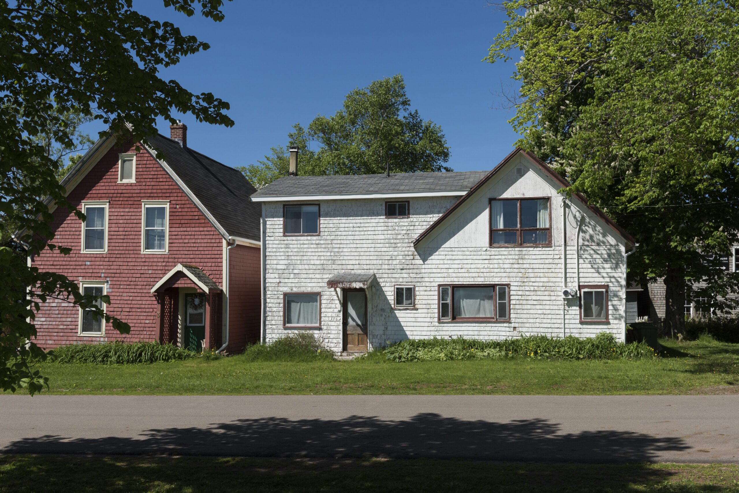 A-frame wooden homes sit side by side on a road. They are surrounded by mature trees but the paint on each is peeling.