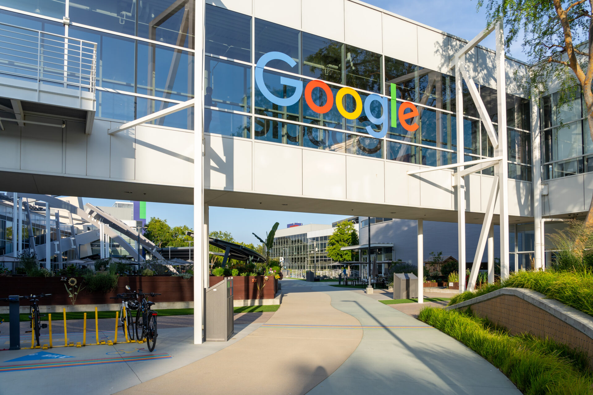 The Google logo adorns a glass-panelled overhead walkway over a concrete path that leads to a couple other low-rise buildings. 