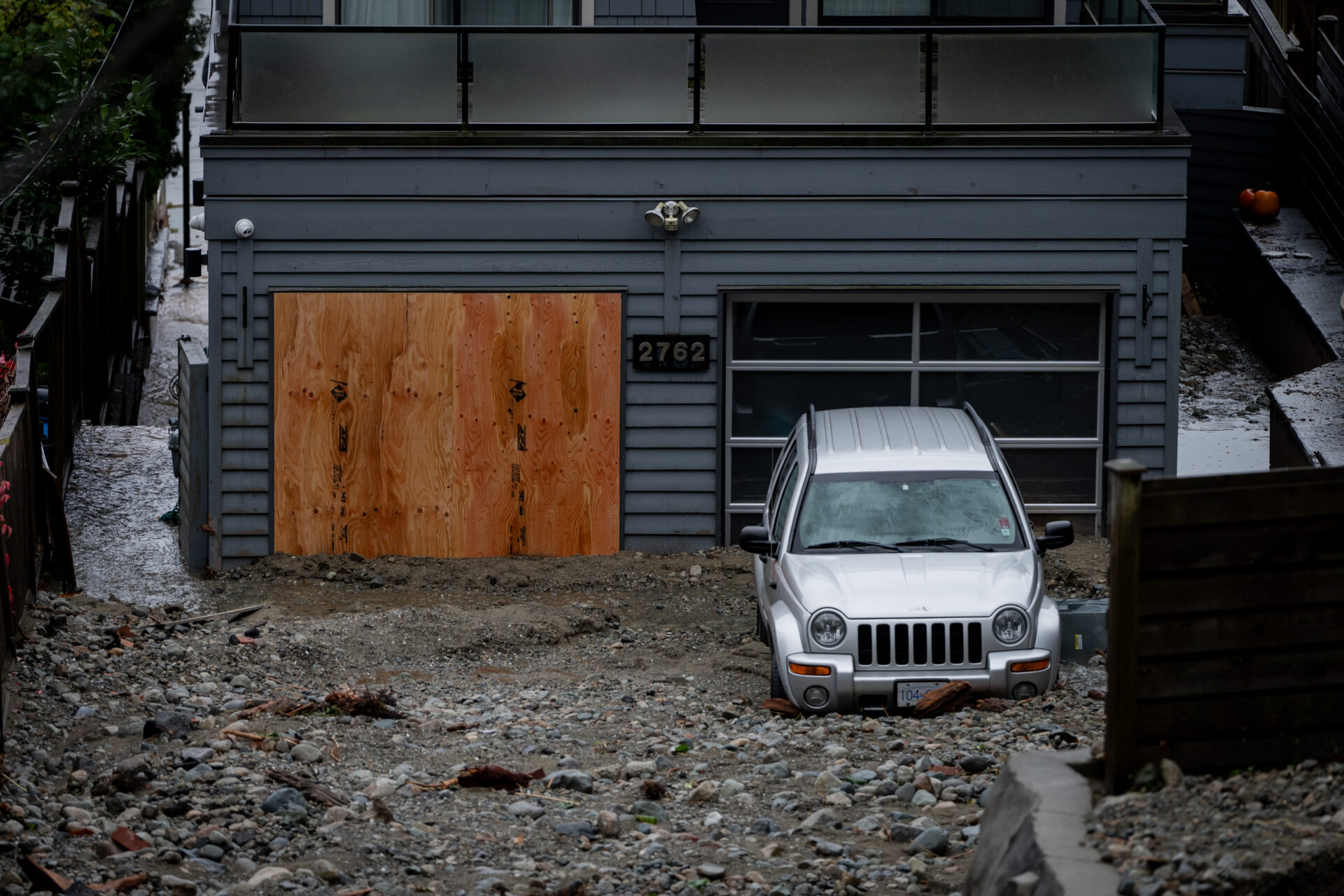 A grey sport-utility vehicle sits on several feet of small rocks, gravel and soil. The door to the home is covered in plywood.