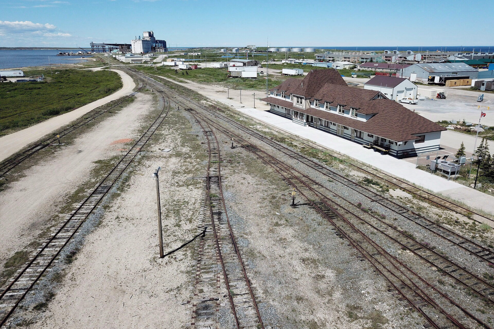 The rail lines are rusted and overgrown as they pass in front of the station, a long white building with a steep pitched roof with wide overhands. The rail lines, seven of them, converge here and run toward a body of water in the distance. The area is industrial, with round storage tanks and warehouses.
