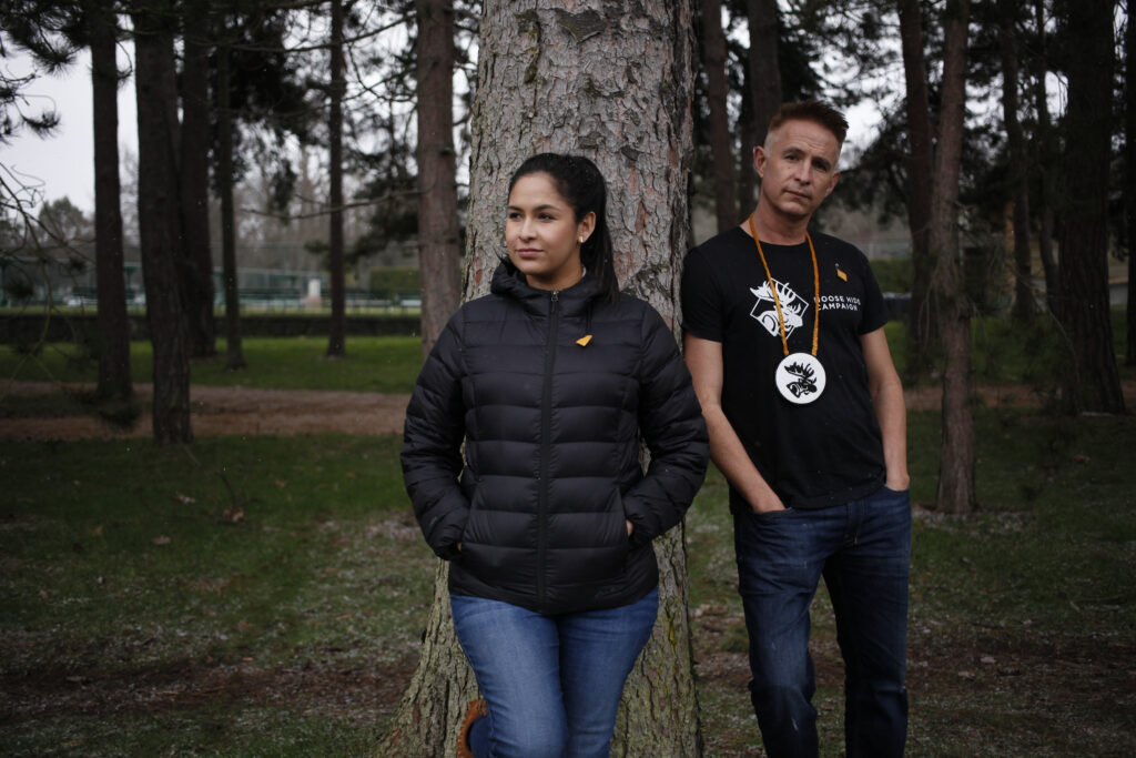The two are both leaning against a wide tree trunk in a park area with other mature trees all around. Raven is in a black puffer jacket and jeans. Paul is in jeans and a black t-shirt.