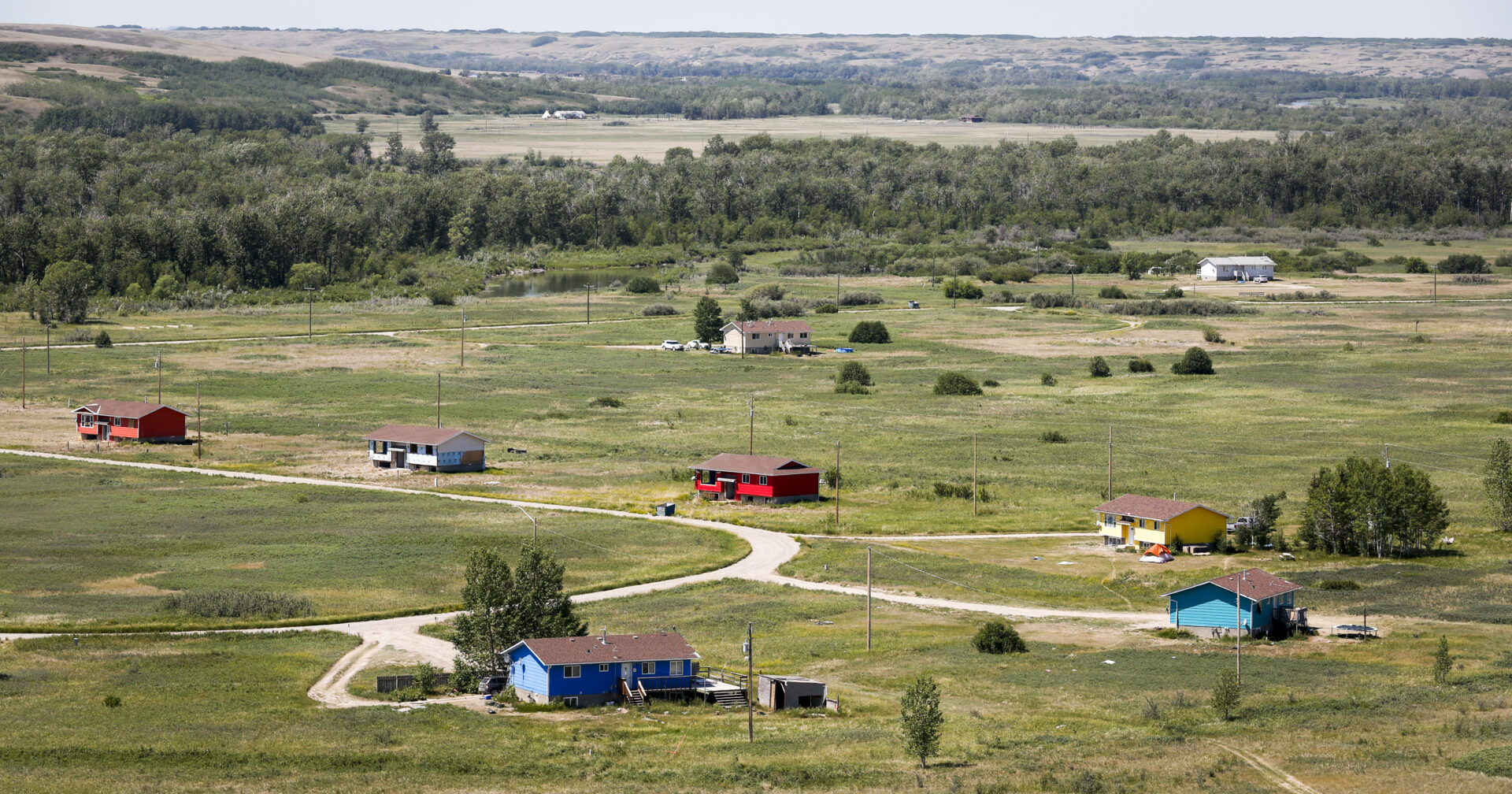 A handful of bungalows sit on a cul-de-sac on a flat, grassy expanse of land bordered by a stand of trees, with fields and rolling hills in the distance. The homes are placed far apart from one another.