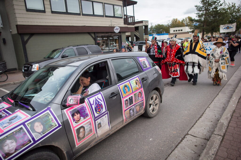 Radek is in tears, with her hand to her face. Behind the car, the people walking are dressed in traditional Indigenous clothing with fringes, straw hats, capes. A few hold signs that mention the Highway of Tears.