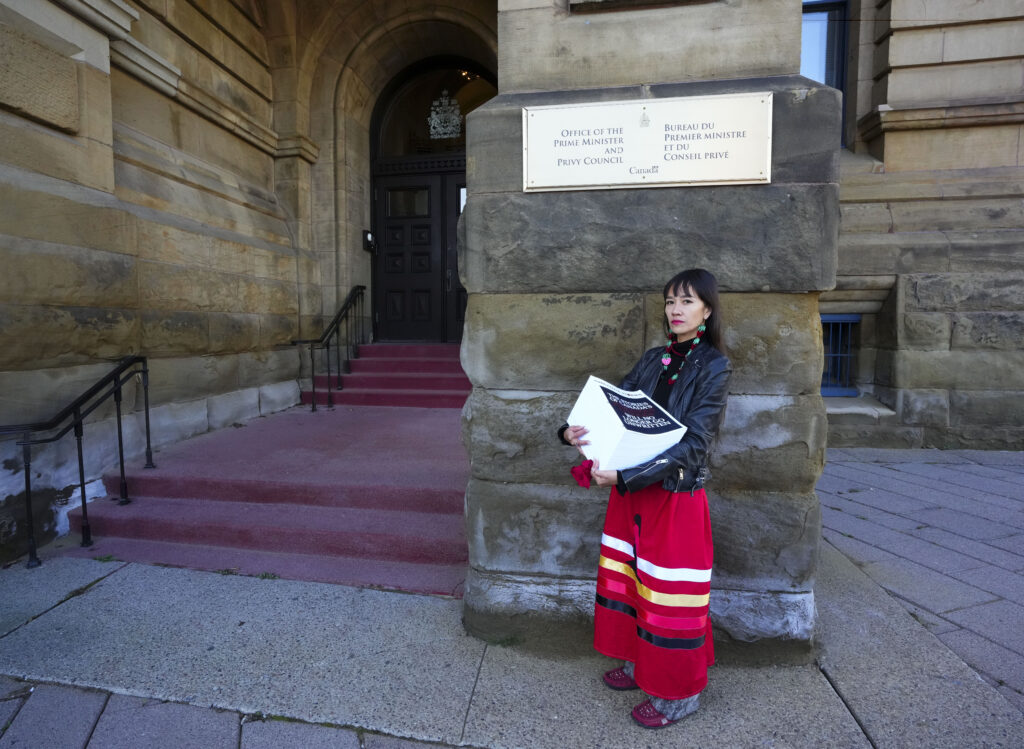 The ânewspaperâ is a tidy, weighty stack of white pages more than a foot thick. Hart is holding it in her arms as she stands in front of the building entrance, which is made of large textured stone pieces. A fading and dusty red carpet covered two sets of shallow stairs leading to the front door, which is arch-shaped in dark wood and a small stained glass window. Hart is wearing a red ribbon skirt, red moccasins with grey fur, a black leather jacket, with a turquoise and pink necklace.