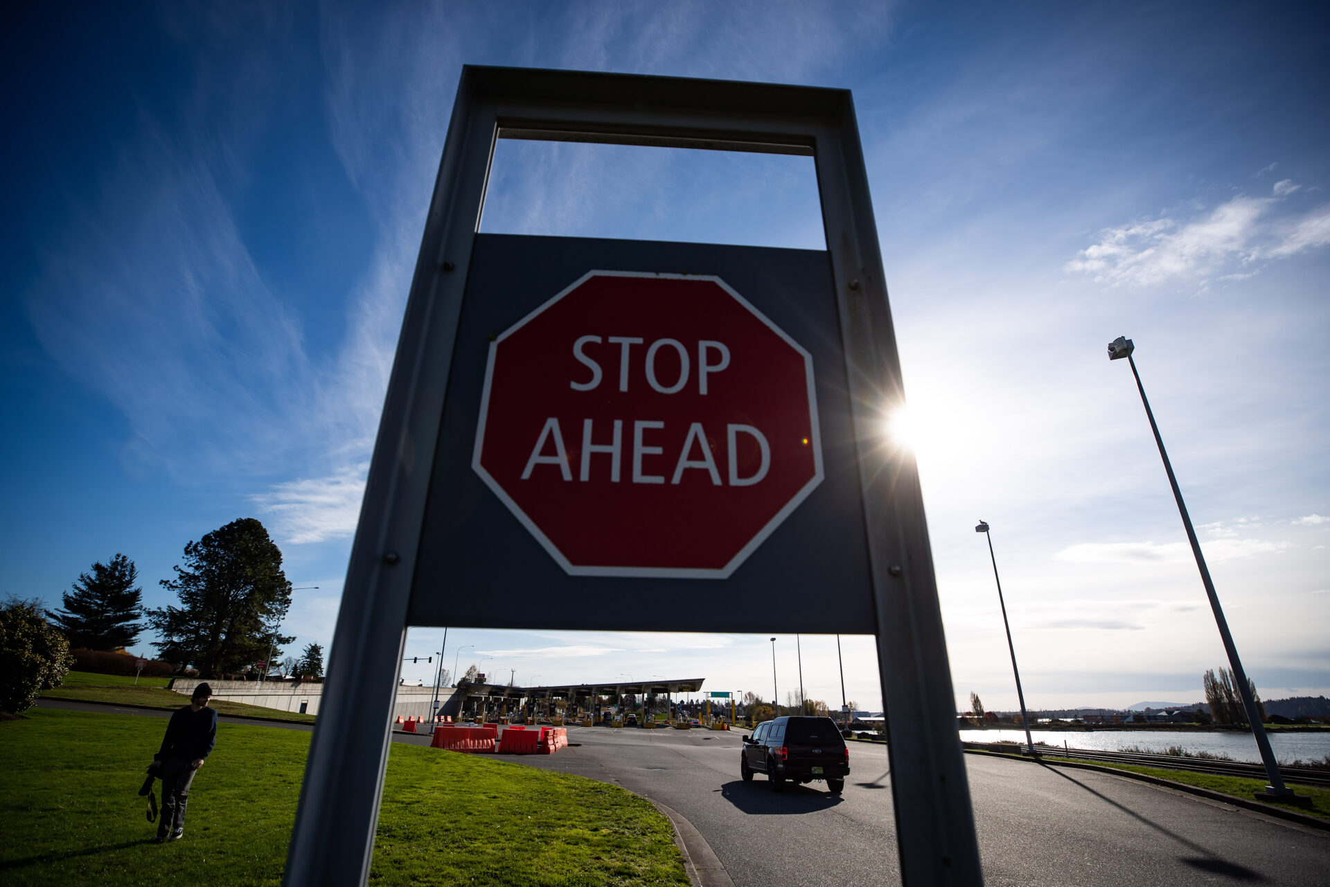 A car passes a stop sign saying “stop ahead” as it approaches the booths. The roadway has a grassy area on one side and water on the other. 